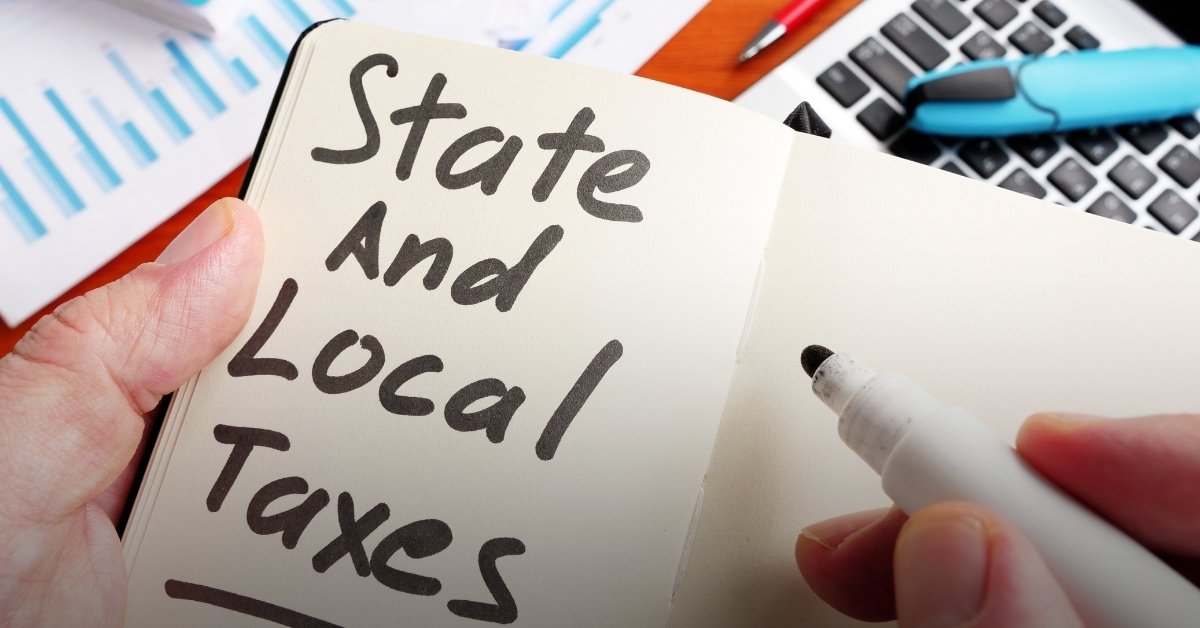 Image of a desk and calculator, with hands holding a pen and notebook with the phrase written: 'State and Local Tax'